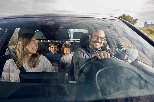 Happy parents and their kids enjoying while going on a trip by car. The view is through glass.